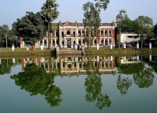 River Cruise To Murapara Zamindar Palace A historical building with a grand facade stands by a tranquil lake, its reflection mirroring the architectural details and surrounding trees.