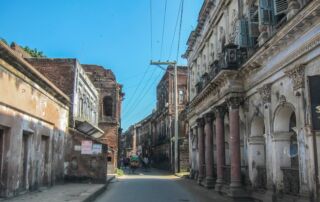 History And Culture In Panam City And Sonargaon A street view with dilapidated buildings on either side and rickshaws in the distance, under a bright blue sky.