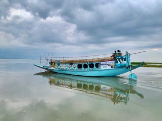 Nikli Haor To Chatir Char Swamp Forest By Boat – Wetlands Exploration A blue boat with colorful decorations floats on calm water under a cloudy sky, with its reflection visible in the water.