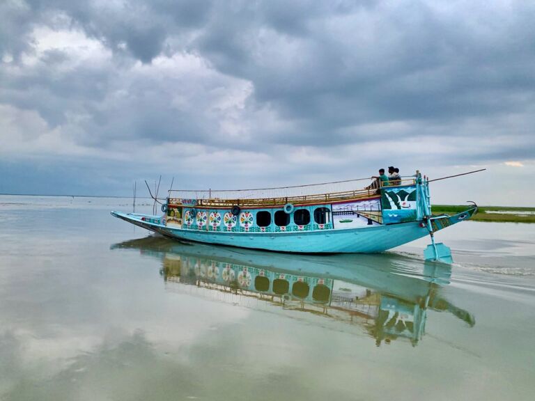 A blue boat with colorful decorations floats on calm water under a cloudy sky, with its reflection visible in the water.