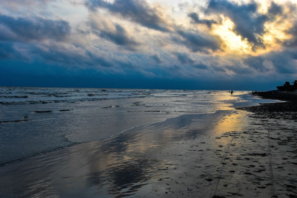 A moody beach scene featuring dark clouds, a reflective wet shoreline, and distant figures, illuminated by a sliver of golden sunlight.
