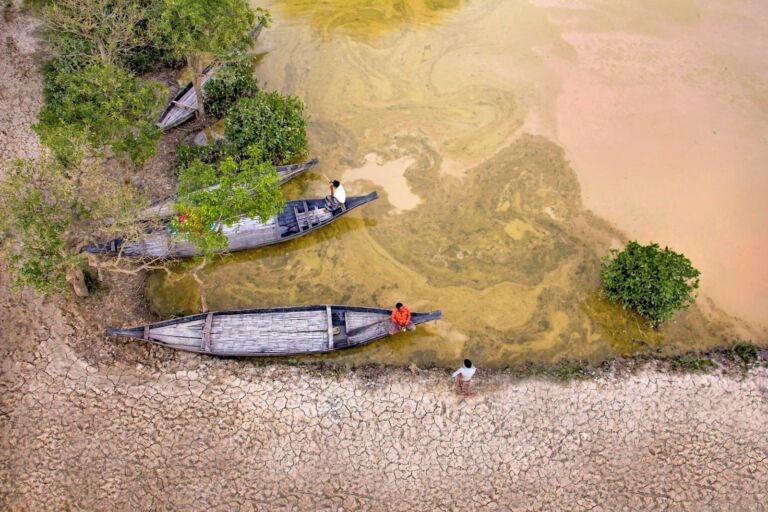 An aerial view shows three narrow boats on a muddy shore, each with a person, amidst a cracked, dry landscape and murky water.