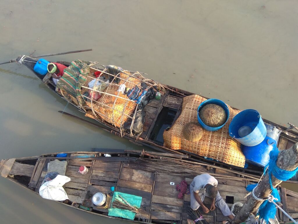 An overhead shot captures two boats floating on a river, one laden with goods and people, the other with a man tending to items on the wooden deck.