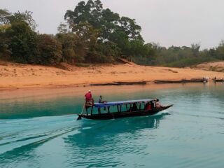 A tour boat carrying passengers travels on a river with turquoise-colored water, with a sandy shore and trees in the background.