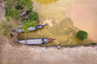 From an aerial perspective, the image shows three narrow boats docked along a cracked, dry shoreline with people and a stagnant, yellowish-brown body of water containing vegetation.