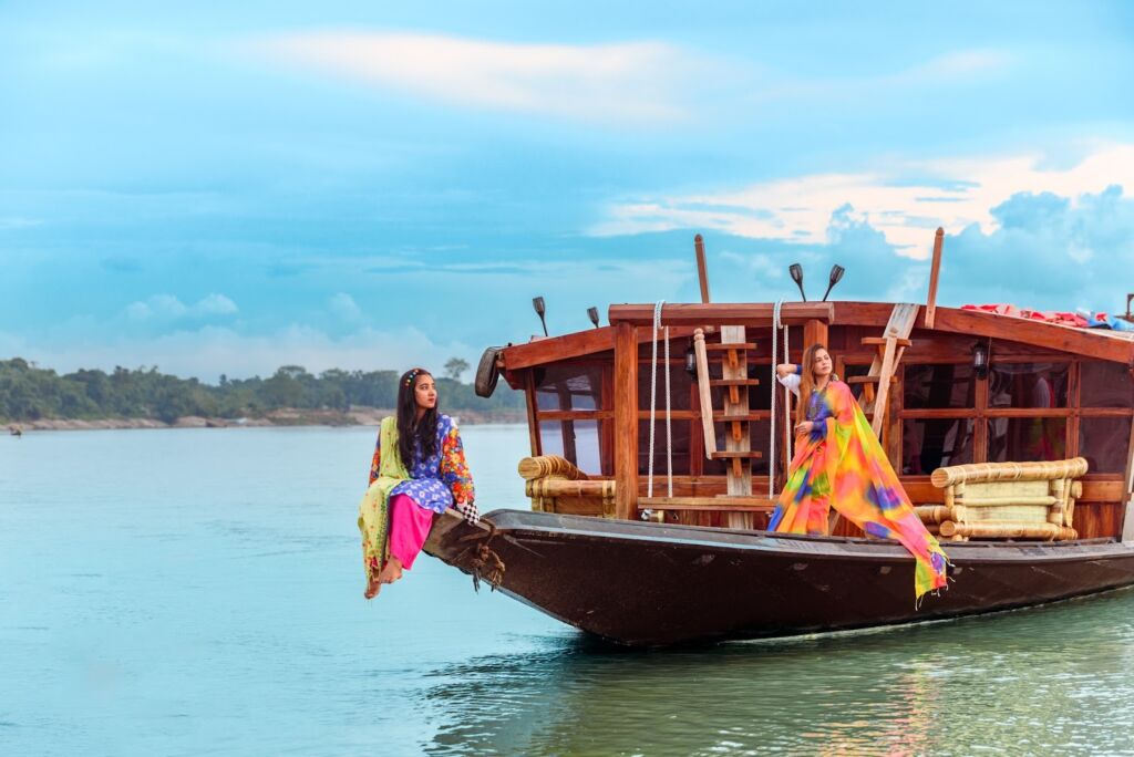 Two women are on a traditional wooden boat on a river. One sits on the bow, the other stands further back near the boat's cabin. Both wear colorful clothing.