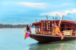 Tanguar Haor Houseboat Experience Two women are on a traditional wooden boat on a river. One sits on the bow, the other stands further back near the boat's cabin. Both wear colorful clothing.