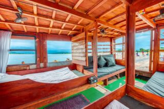 An interior view of a wooden houseboat with beds, pillows, and windows overlooking a lake with people swimming and a sandy beach.