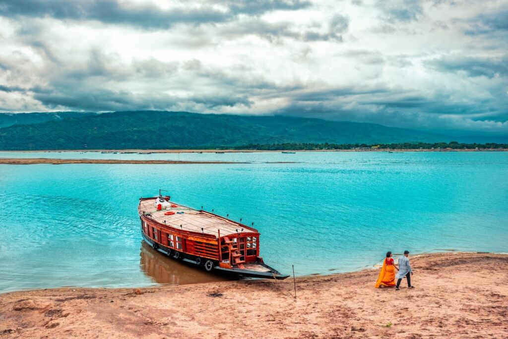 A couple walks along a sandy beach beside a calm turquoise lake, with a large wooden boat docked nearby, against a backdrop of green hills and a cloudy sky.