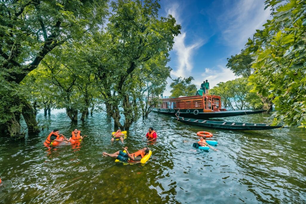 People swim with life jackets in a lake with submerged trees, near a boat, under a blue sky.