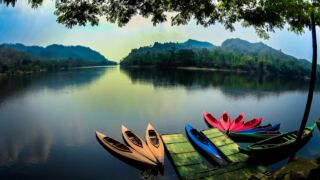 Kaptai Lake And Sajek Valley Waterways Tour A scenic view of a calm river surrounded by green mountains, with colorful canoes docked at a wooden platform.