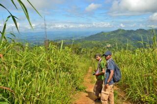 Keokradong Hiking Tour From Chittagong Two young men stand on a narrow path, flanked by tall green grass, with a scenic view of rolling green hills and a partly cloudy sky in the background.
