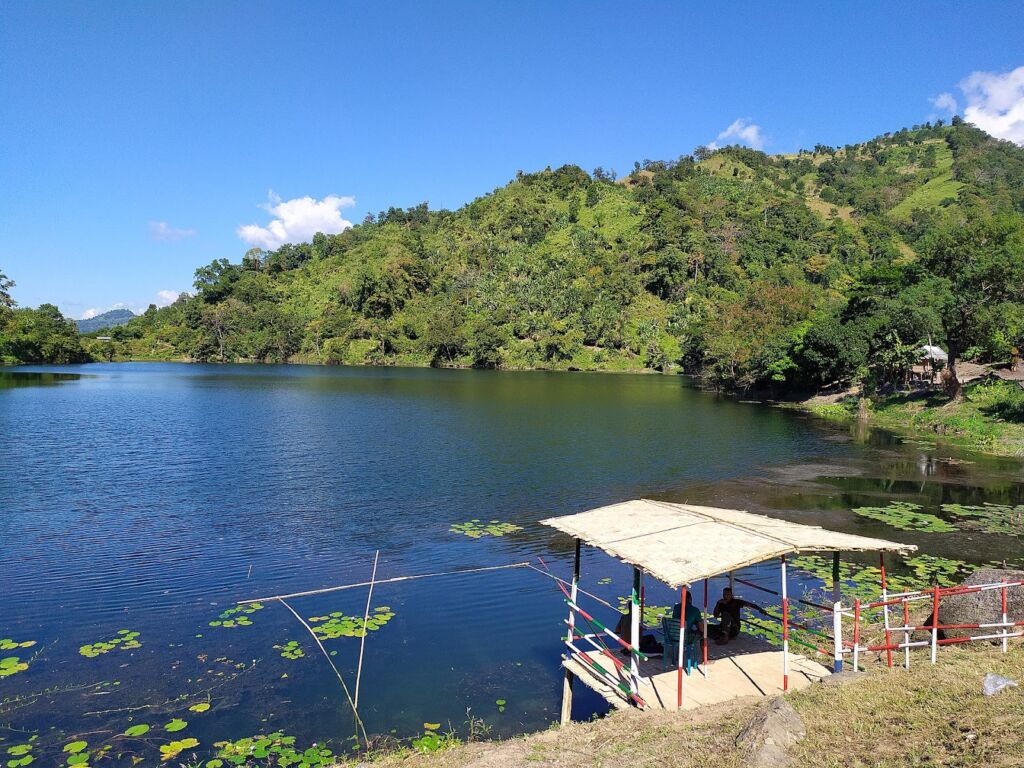 A serene lake surrounded by lush green hills with a small boathouse in the foreground, under a clear blue sky.