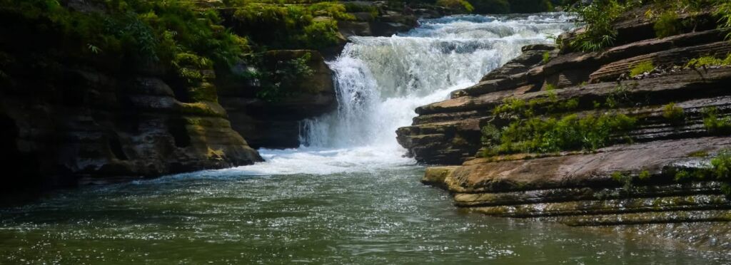 A scenic view of a waterfall cascading down layered rocks into a river, surrounded by lush green vegetation.