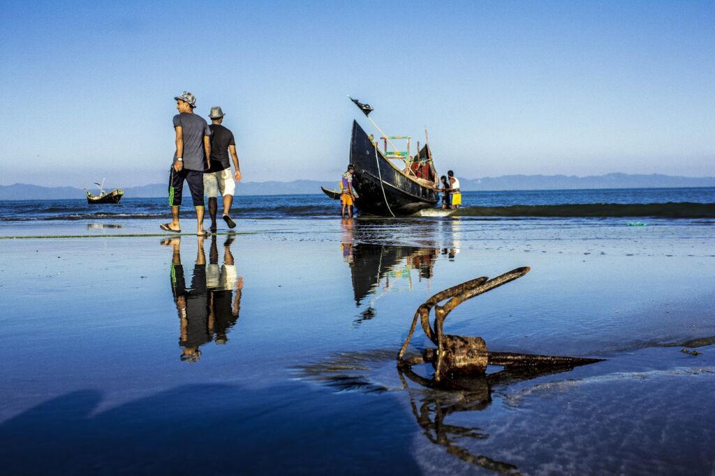 Two men walk along a reflective beach towards a grounded fishing boat, with a rusty anchor in the foreground and other boats visible on the horizon.