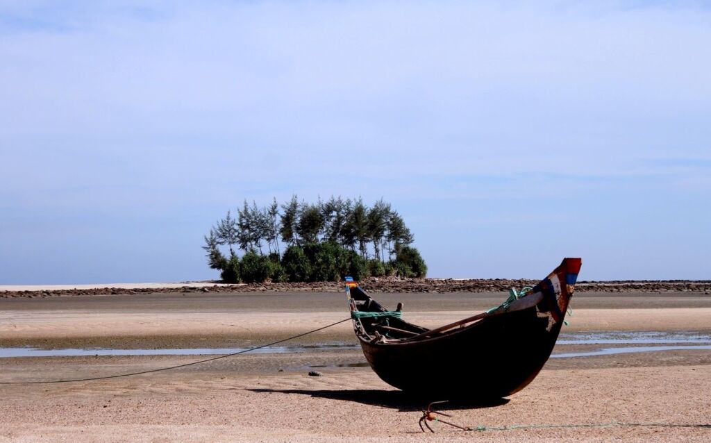 A wooden boat sits beached on a sandy shore with a small, tree-covered island in the background under a pale blue sky.