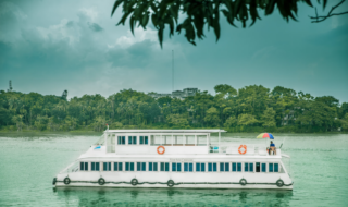 Exploring Bangladesh’s Wetlands: Boat ride from Nikli Haor To Chatir Char Swamp Forest A white two-story boat with blue windows floats on a calm green lake under a cloudy sky with lush green trees on the shore.