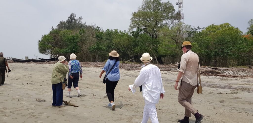A group of people are walking on a beach towards a line of trees and a tower on a cloudy day.