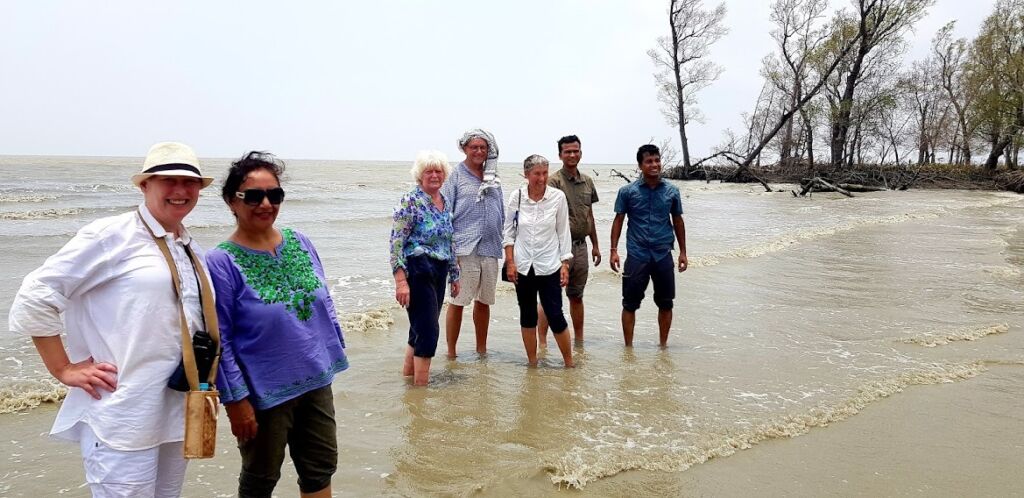 A group of seven people stand in shallow water at a beach with trees in the background.