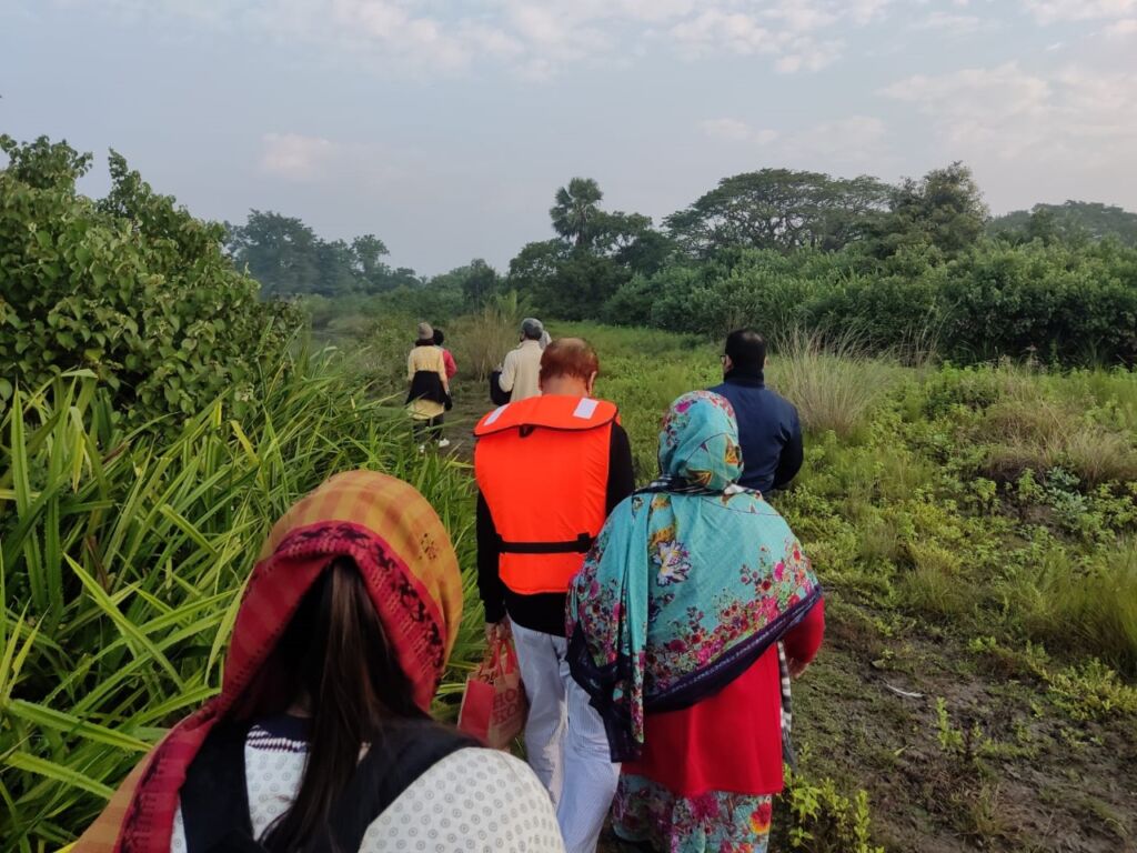 A group of people is walking on a dirt path surrounded by lush greenery. One person is wearing a life jacket.