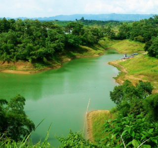Explore Bangladesh’s unique attraction: The Kaptai lake A lush green landscape features a river winding through dense foliage, with boats docked on the grassy bank under a partly cloudy sky.