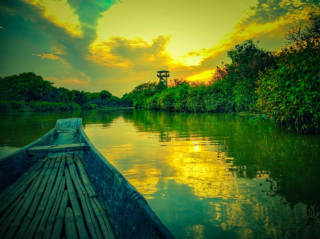 Ratargul and Bholaganj Boat Cruise: Scenic Nature Exploration A serene view from a boat on a river at sunset, with lush green vegetation and a distant tower.