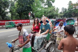 Full-Day Dhaka Exploration Tour Two women are riding in a decorated rickshaw pulled by a man in a street in Bangladesh, with other rickshaws and people nearby.