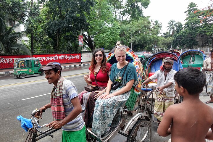 Two women are riding in a decorated rickshaw pulled by a man in a street in Bangladesh, with other rickshaws and people nearby.