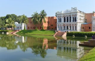 Explore the Best of Dhaka & Sonargaon: A Full-Day Guided Tour A scenic view of a palace and buildings reflected in a pond with palm trees and lush greenery under a clear blue sky.