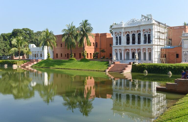 A scenic view of a palace and buildings reflected in a pond with palm trees and lush greenery under a clear blue sky.